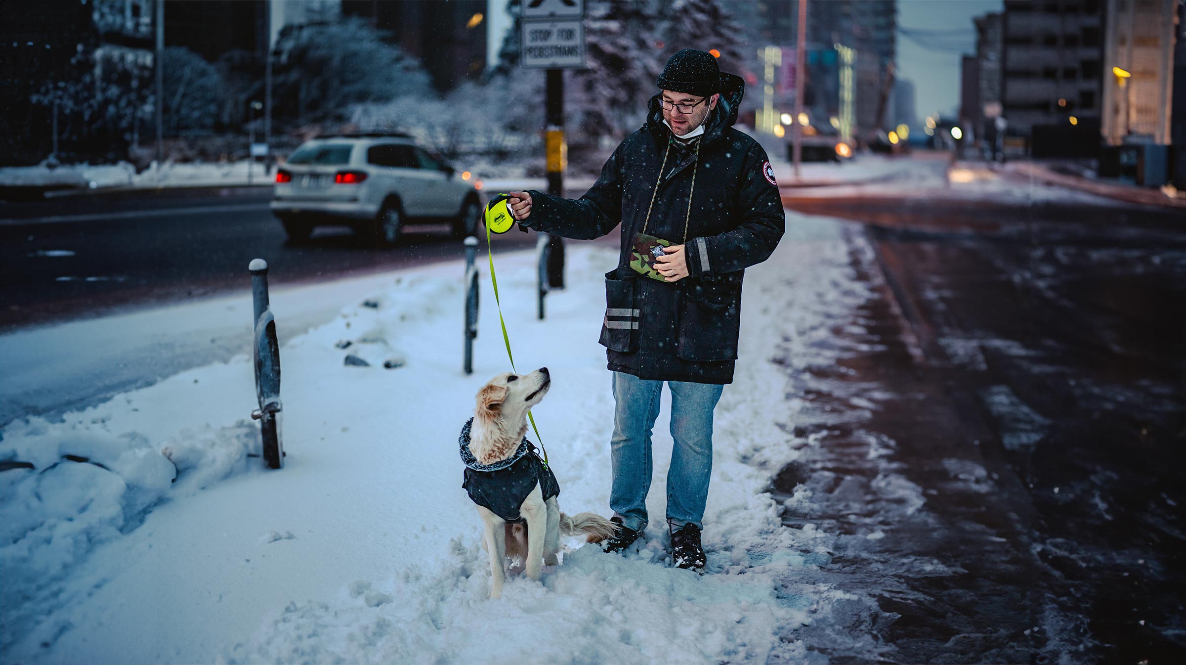 Man wearing holding a retractable leash, standing in the snow with a golden retriever looking up at him